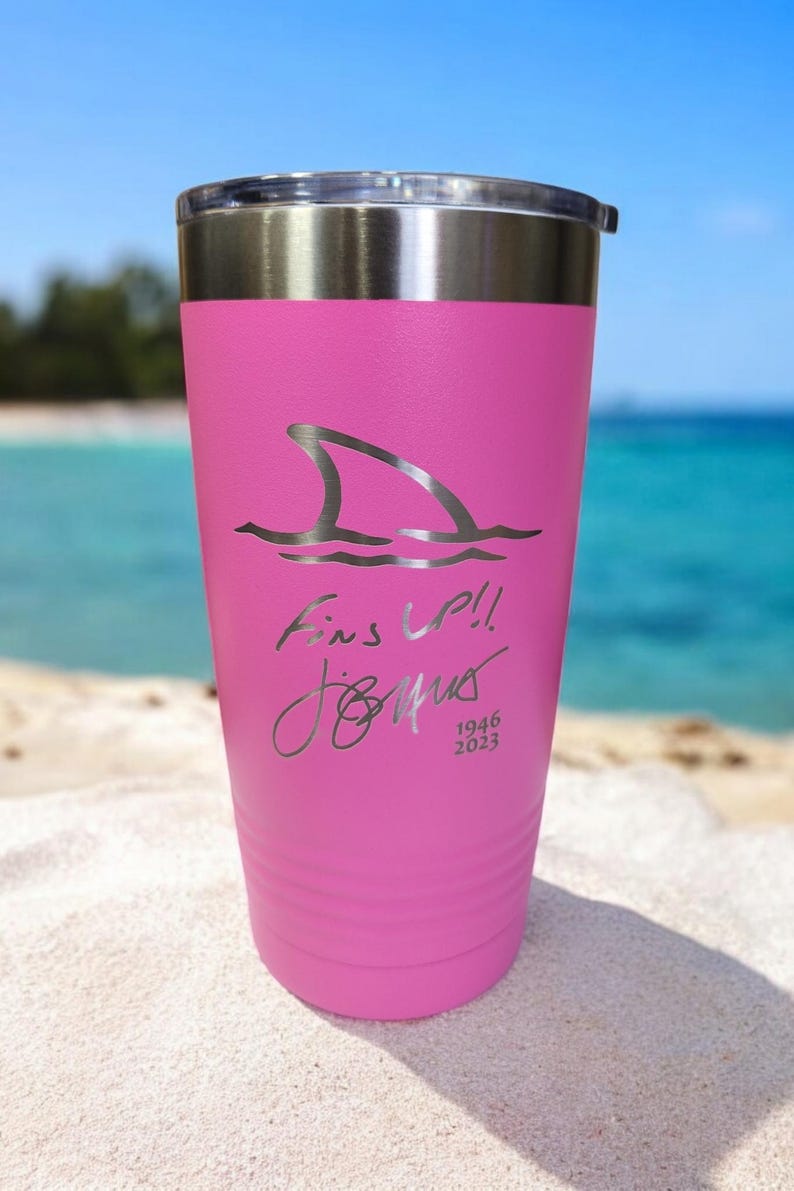 May include: A pink insulated tumbler with a silver lid. The tumbler features a shark fin graphic and the text "Fins Up!!" with a signature and the dates 1946 and 2023. The background shows a beach and ocean.