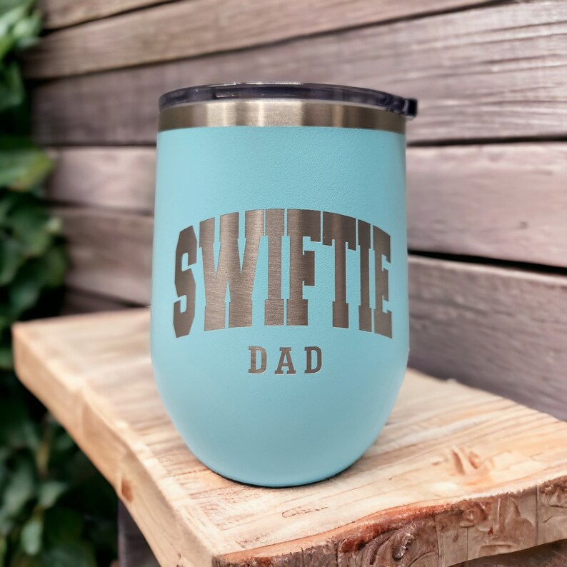 May include: A light blue stainless steel tumbler with a silver inscription that reads "Swiftie Dad".