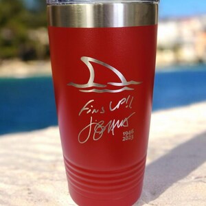 May include: A red insulated tumbler with a silver lid. The tumbler features a shark fin graphic above the words "Fins Up!!" and a signature, with the dates "1946 2023" below. The tumbler is on a sandy surface.