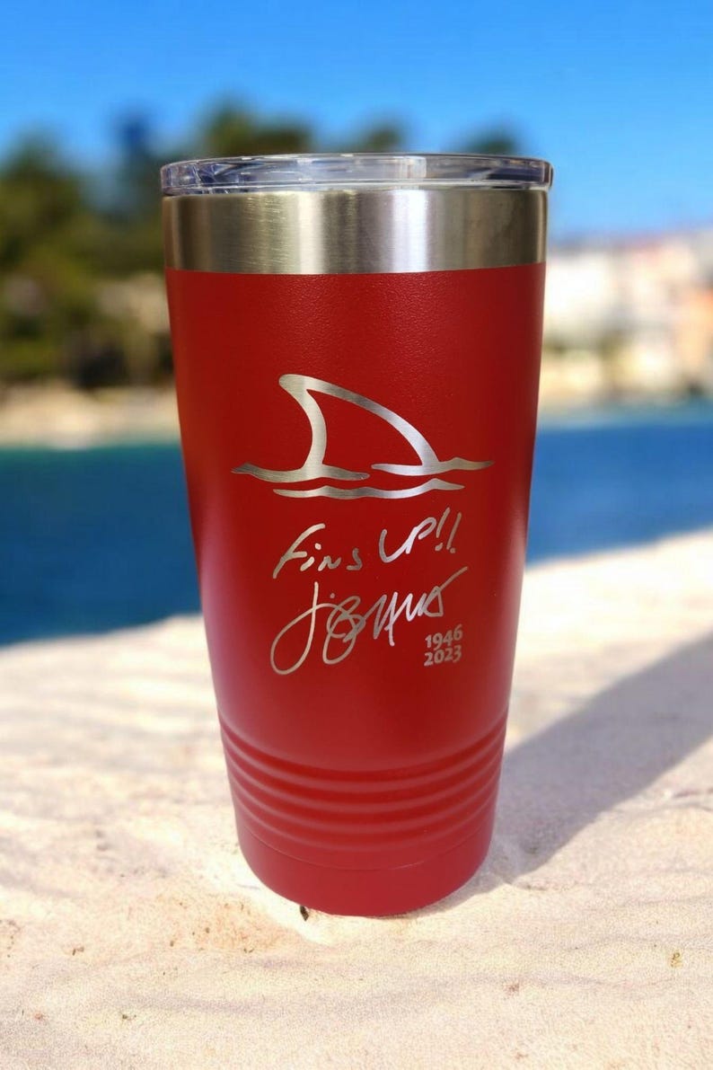May include: A red insulated tumbler with a silver lid. The tumbler features a shark fin graphic above the words "Fins Up!!" and a signature, with the dates "1946 2023" below. The tumbler is on a sandy surface.