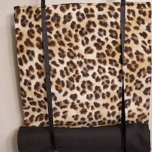 May include: A black and brown leopard print fabric roller shade with black fabric ties. The shade is rolled up and hanging from two black hooks.