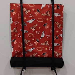 May include: A red fabric roller shade with a cowboy boot and lasso pattern. The shade is mounted to a wall with two white hooks and has black straps.