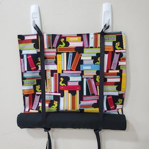 May include: A black fabric book roll with a colorful book print featuring titles like "Chemistry", "English", "History", and "Dictionary". The roll is attached to two white hooks on a wall with black straps.