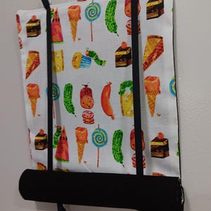 May include: A white fabric window shade featuring a colorful pattern of various sweets, including ice cream cones, lollipops, cakes, and a caterpillar. The shade has a black bottom and black straps for hanging.