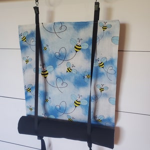 May include: A blue and white fabric roller shade with a bee pattern. The shade is hanging from a black rod with two black straps.