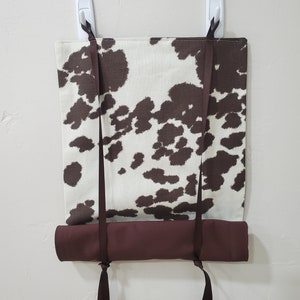 May include: A brown and white cow print fabric roller shade with brown fabric ties. The shade is mounted on a white wall with two white plastic hooks.