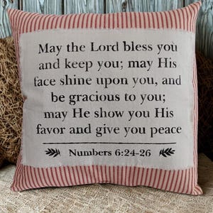 May include: Square decorative pillow with a white center and a red and white striped border. The center features a printed religious quote from Numbers 6:24-26 in black text. The pillow is on a neutral-toned surface.
