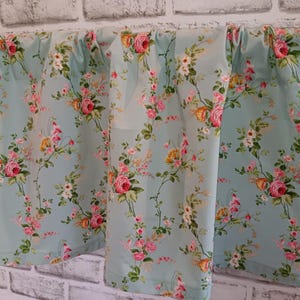 May include: Light blue floral curtains with a repeating pattern of pink and yellow roses and white flowers. The curtains are hanging in front of a white brick wall. The floral design is a classic, vintage style.