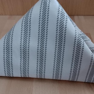 May include: Folded white cloth napkin with a gray striped pattern. The napkin is folded into a triangle shape, with the pattern running diagonally across the surface. The napkin is resting on a wooden surface.