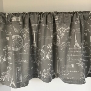May include: Gray and white valance with a Parisian theme. The valance features a repeating pattern of bicycles, Eiffel Towers, clocks, and French text. The valance is made of a soft, textured fabric.