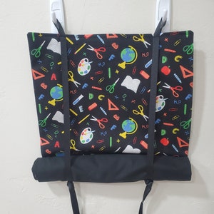 May include: Black fabric wall hanging with a school-themed pattern featuring colorful images of pencils, scissors, rulers, globes, and apples. The fabric is attached to two white hooks with black straps.