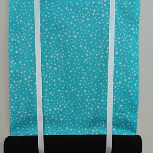 Puede incluir: Una cortina de tela turquesa con un estampado de lunares blancos. La cortina tiene una parte inferior enrollada negra y está sujeta por dos cintas blancas. La cortina está colgada en una pared blanca.