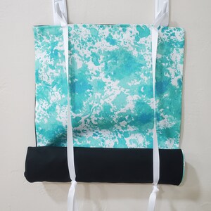 May include: A teal and white patterned yoga mat with black trim is rolled up and hanging on a wall with white hooks. The mat has white straps that are tied together at the bottom.