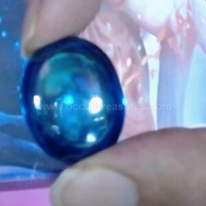 May include: A smooth, oval-shaped blue gemstone with a polished, reflective surface. The stone displays a deep, translucent blue color with lighter highlights. The text www.occultreasures.com is visible across the center of the gemstone.