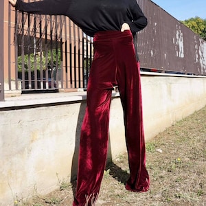 May include: A woman wearing a black long-sleeved shirt and wide-leg, burgundy velvet pants. She is standing in front of a metal fence.