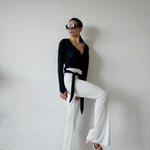 May include: A woman wearing a black long-sleeved wrap top with a black tie belt and white flared pants. She is wearing sunglasses and has her hair pulled back.