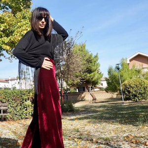 May include: A woman wearing a black long-sleeved shirt with fringe detail and burgundy velvet wide-leg pants. She is standing in a park with trees and grass in the background.