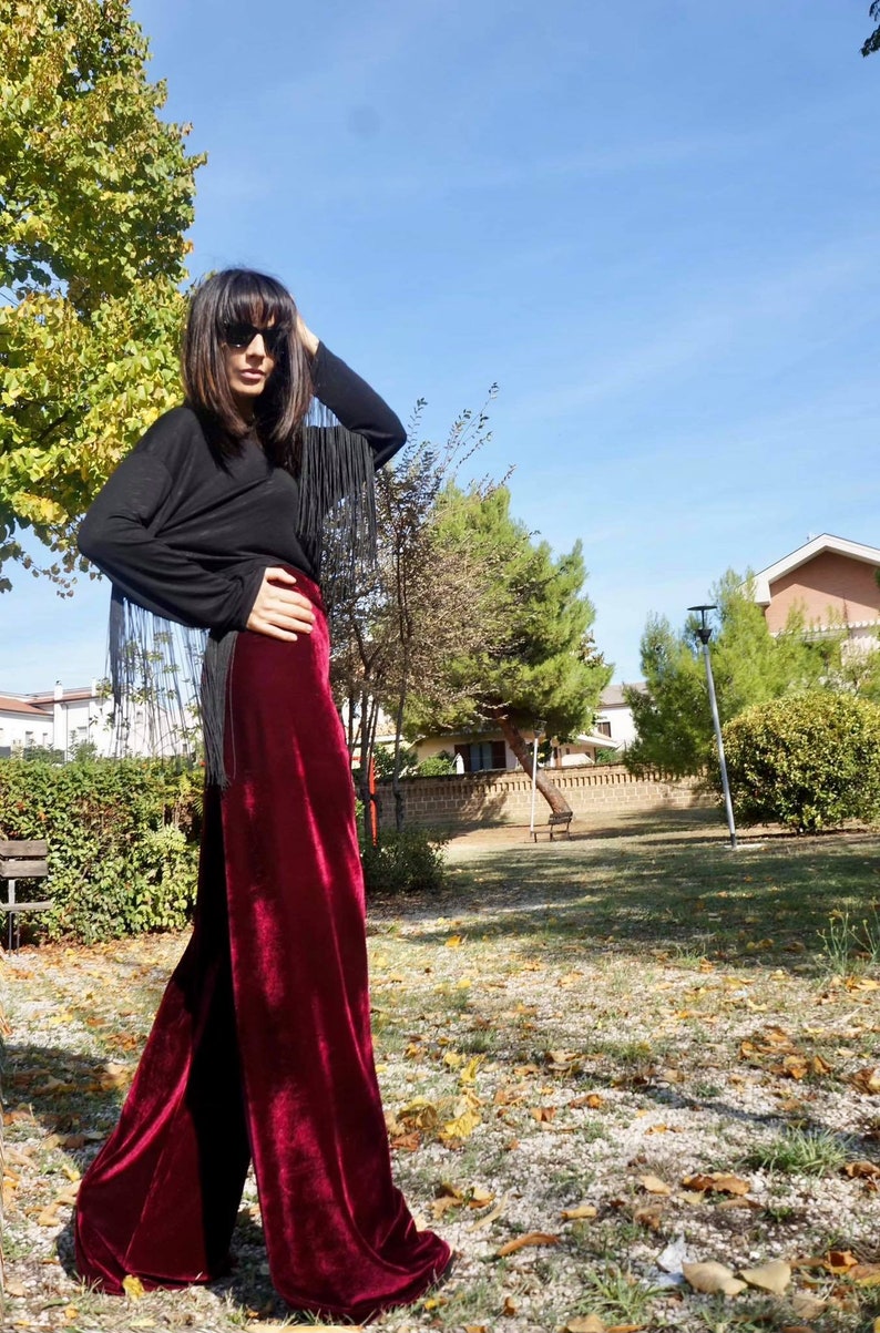 May include: A woman wearing a black long-sleeved shirt with fringe detail and burgundy velvet wide-leg pants. She is standing in a park with trees and grass in the background.