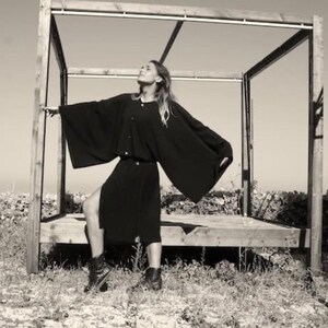 May include: A woman wearing a long, black, flowing dress with a button-up front. She is standing in front of a wooden structure with a slatted roof.