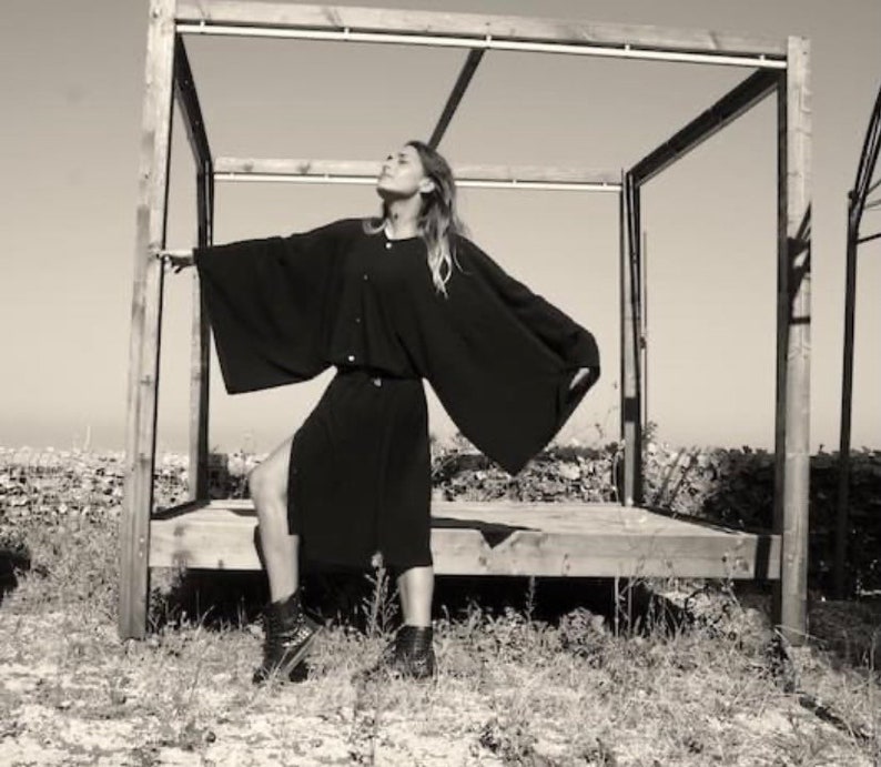 May include: A woman wearing a long, black, flowing dress with a button-up front. She is standing in front of a wooden structure with a slatted roof.