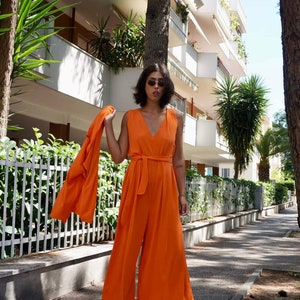 Puede incluir: Un mono naranja con una chaqueta a juego. El mono sin mangas tiene cuello en V y cintur&oacute;n anudado. La chaqueta se sostiene sobre el brazo. La persona lleva gafas de sol y sandalias. El fondo incluye una valla blanca y un edificio.
