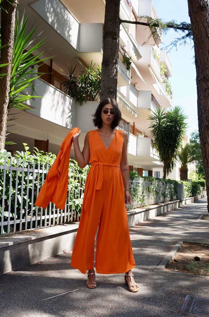 Puede incluir: Un mono naranja con una chaqueta a juego. El mono sin mangas tiene cuello en V y cintur&oacute;n anudado. La chaqueta se sostiene sobre el brazo. La persona lleva gafas de sol y sandalias. El fondo incluye una valla blanca y un edificio.