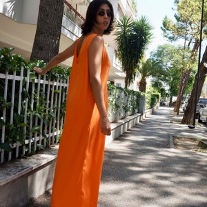 Puede incluir: Un vestido largo naranja sin mangas. El vestido tiene un dise&ntilde;o sencillo y fluido con un escote en V profundo en la espalda. La persona lleva gafas de sol y sandalias. El vestido es de un color vibrante y liso, adecuado para el clima c&aacute;lido.