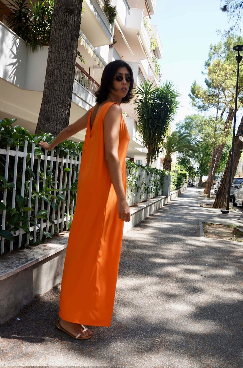 Puede incluir: Un vestido largo naranja sin mangas. El vestido tiene un dise&ntilde;o sencillo y fluido con un escote en V profundo en la espalda. La persona lleva gafas de sol y sandalias. El vestido es de un color vibrante y liso, adecuado para el clima c&aacute;lido.