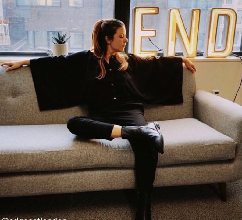 May include: A woman wearing a black shirt and black pants sits on a gray couch. She is looking to the side. Behind her is a neon sign that says "END".