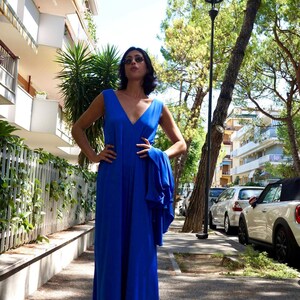 Puede incluir: Un vestido largo maxi sin mangas de color azul real con cuello en V. El vestido llega hasta el suelo y parece estar hecho de una tela fluida. La persona lleva gafas de sol y sandalias. El fondo incluye edificios, &aacute;rboles y coches aparcados.