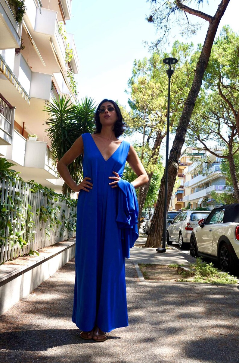Puede incluir: Un vestido largo maxi sin mangas de color azul real con cuello en V. El vestido llega hasta el suelo y parece estar hecho de una tela fluida. La persona lleva gafas de sol y sandalias. El fondo incluye edificios, &aacute;rboles y coches aparcados.