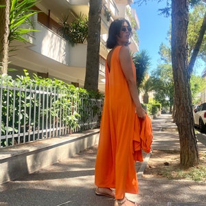 Puede incluir: Un mono naranja sin mangas con perneras anchas. La persona lleva sandalias doradas y gafas de sol. El conjunto se combina con una chaqueta naranja a juego. El fondo incluye &aacute;rboles, una acera y un edificio.