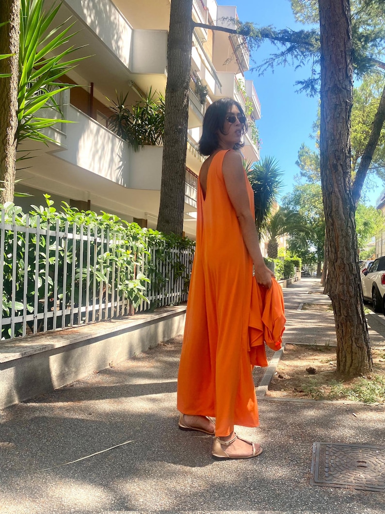 Puede incluir: Un mono naranja sin mangas con perneras anchas. La persona lleva sandalias doradas y gafas de sol. El conjunto se combina con una chaqueta naranja a juego. El fondo incluye &aacute;rboles, una acera y un edificio.