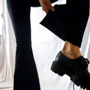 Puede incluir: Una persona con pantalones negros y una camisa blanca, con un tatuaje en el tobillo que dice "I know life". Lleva zapatos negros con suela gruesa.