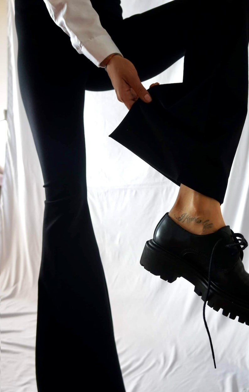 Puede incluir: Una persona con pantalones negros y una camisa blanca, con un tatuaje en el tobillo que dice "I know life". Lleva zapatos negros con suela gruesa.