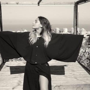 Peut inclure: Une femme portant une chemise noire à manches longues avec un col large et des manches longues. Elle se tient dans une porte avec une vue sur un horizon urbain en arrière-plan.