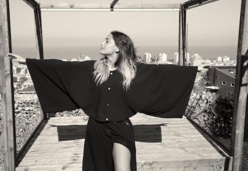 May include: A woman wearing a black long-sleeved shirt with a wide collar and long sleeves. She is standing in a doorway with a view of a city skyline in the background.