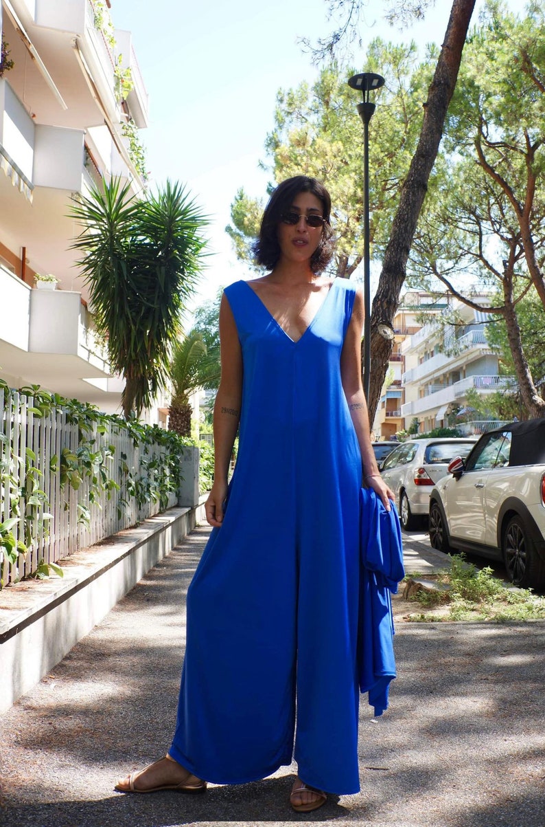 Puede incluir: Mono sin mangas azul real con dise&ntilde;o de pernera ancha y cuello en V. La modelo lleva gafas de sol y sostiene una chaqueta azul a juego. La foto est&aacute; tomada al aire libre en un d&iacute;a soleado.