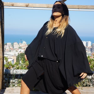 May include: A woman wearing a black, long-sleeved, flowing dress with a button-up front. She is standing on a wooden platform with a view of a city and ocean in the background.