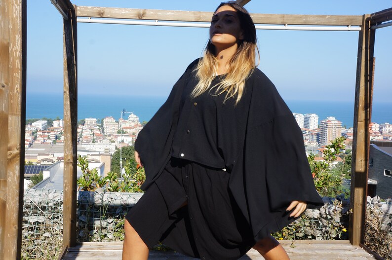 May include: A woman wearing a black, long-sleeved, flowing dress with a button-up front. She is standing on a wooden platform with a view of a city and ocean in the background.