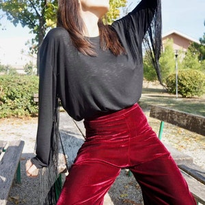 May include: A woman wearing a black long-sleeved shirt with fringe detailing on the sleeves and burgundy velvet wide-leg pants.