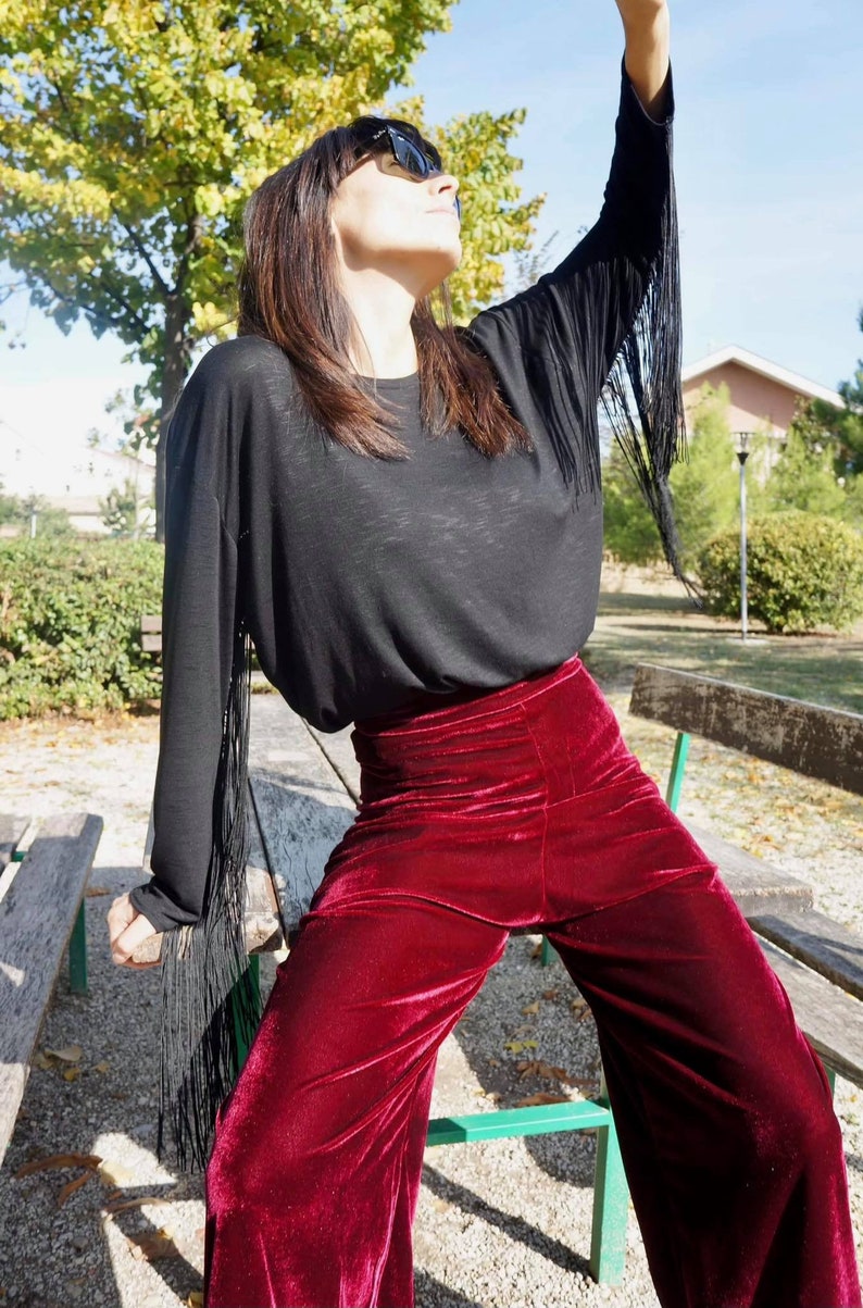 May include: A woman wearing a black long-sleeved shirt with fringe detailing on the sleeves and burgundy velvet wide-leg pants.