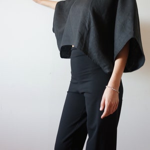 May include: A black linen top with a wide, boxy silhouette and short sleeves. The top is cropped and has a slightly uneven hemline. The model is wearing black pants.