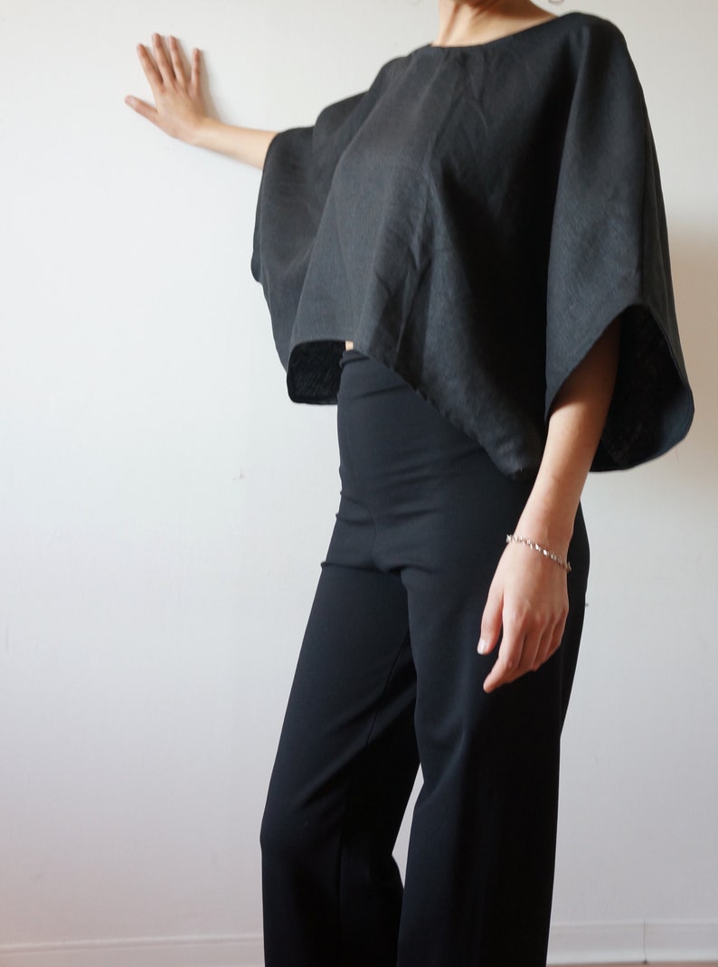 May include: A black linen top with a wide, boxy silhouette and short sleeves. The top is cropped and has a slightly uneven hemline. The model is wearing black pants.