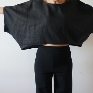 May include: A black linen top with a wide, flowing silhouette. The top is cropped and has a relaxed fit. The model is wearing black pants.