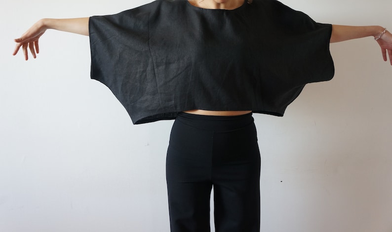 May include: A black linen top with a wide, flowing silhouette. The top is cropped and has a relaxed fit. The model is wearing black pants.