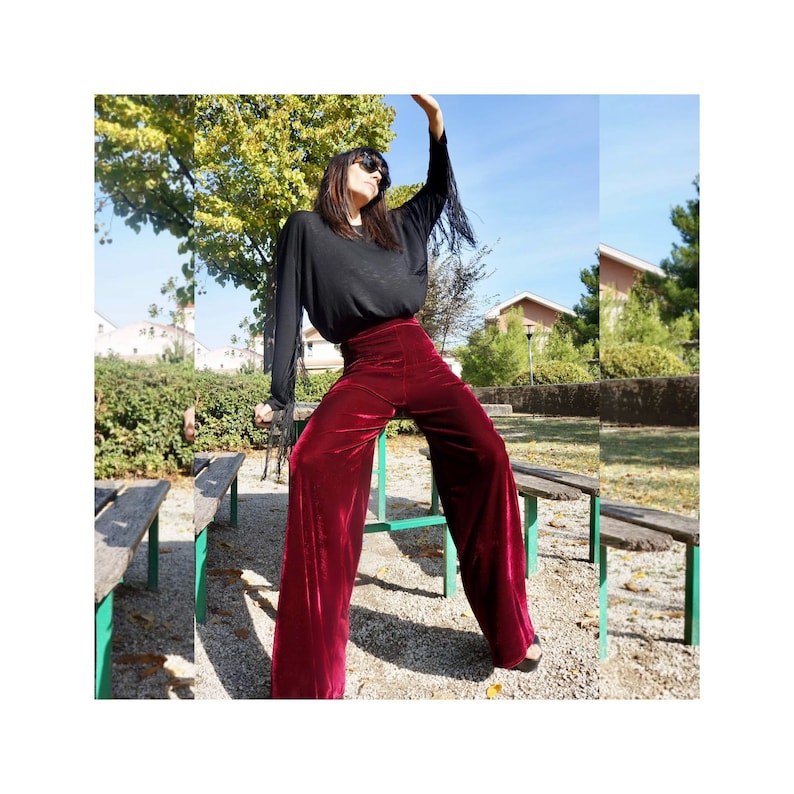 May include: A woman wearing a black long-sleeved shirt with fringe detail and wide-leg, burgundy velvet pants. She is standing on a wooden bench in a park setting.