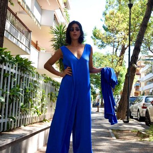 Puede incluir: Un mono azul real sin mangas con un dise&ntilde;o de pierna ancha y un escote en V profundo. El atuendo se combina con una chaqueta azul a juego. La persona lleva gafas de sol y sandalias. El fondo incluye edificios y &aacute;rboles.