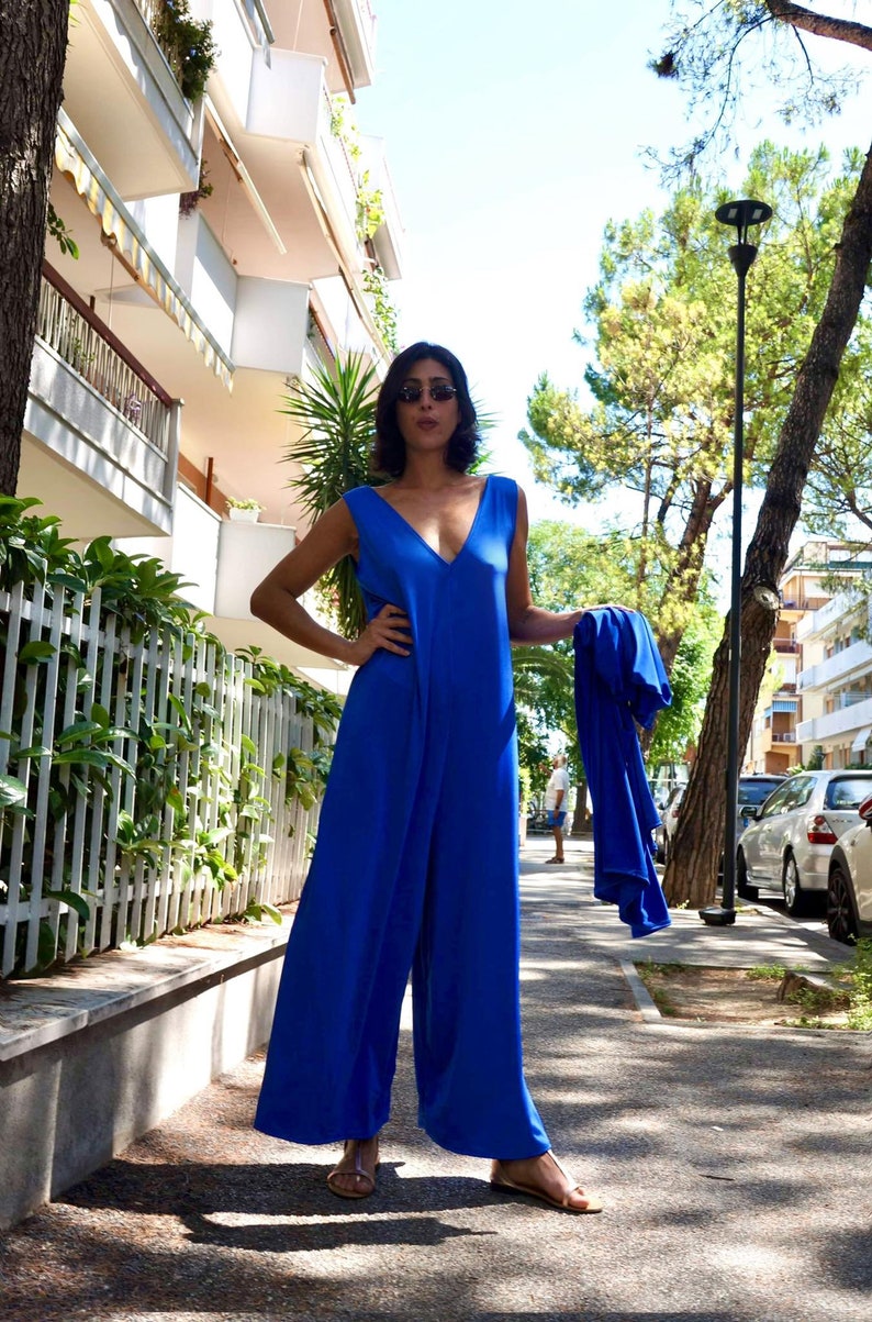 Puede incluir: Un mono azul real sin mangas con un dise&ntilde;o de pierna ancha y un escote en V profundo. El atuendo se combina con una chaqueta azul a juego. La persona lleva gafas de sol y sandalias. El fondo incluye edificios y &aacute;rboles.