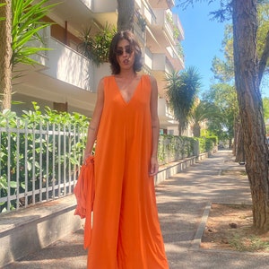 Puede incluir: Un mono naranja sin mangas con un dise&ntilde;o de pierna ancha. El mono tiene un escote en V profundo y se combina con tela naranja a juego. La persona lleva gafas de sol y sandalias. El fondo incluye &aacute;rboles, un edificio y una acera.